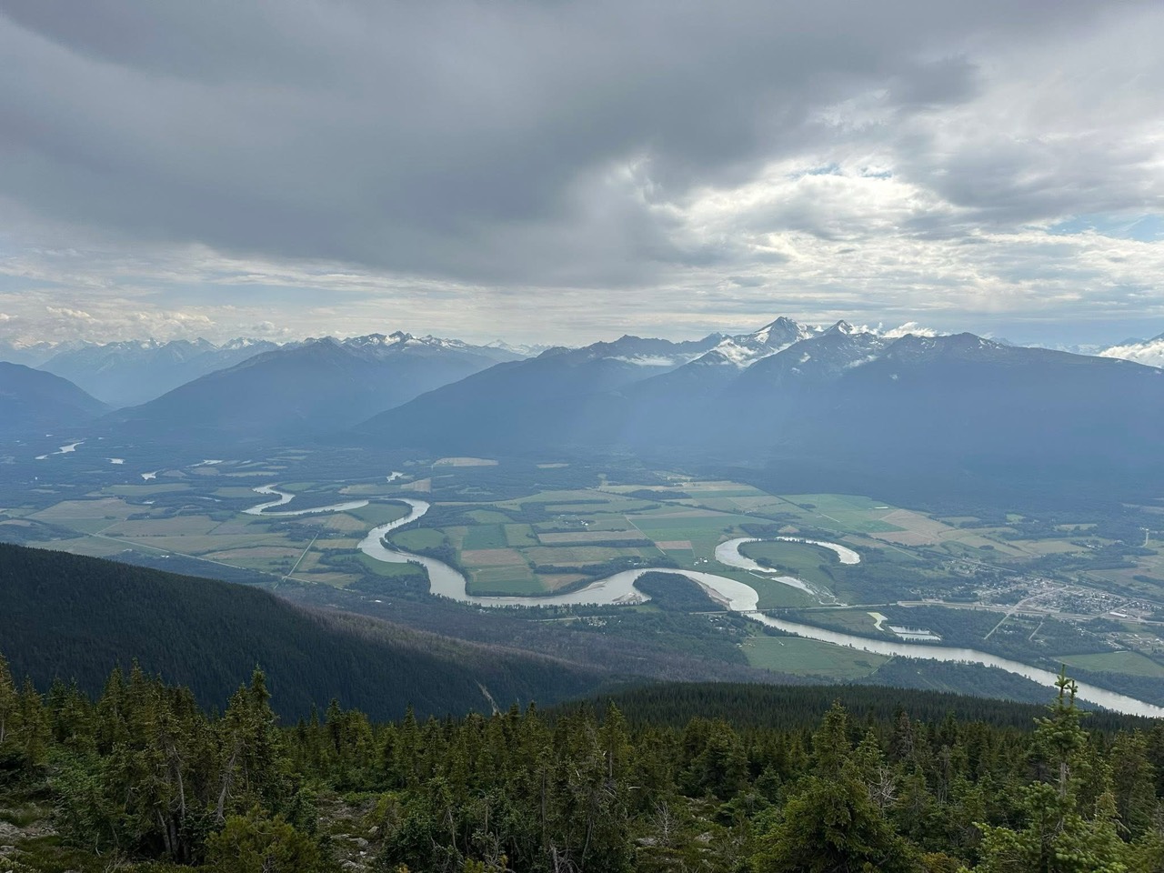 A winding river through green fields below forested slopes and distant mountain ridges, viewed from McBride Peak